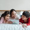 cheerful ethnic mother watching video via tablet with kids on bed